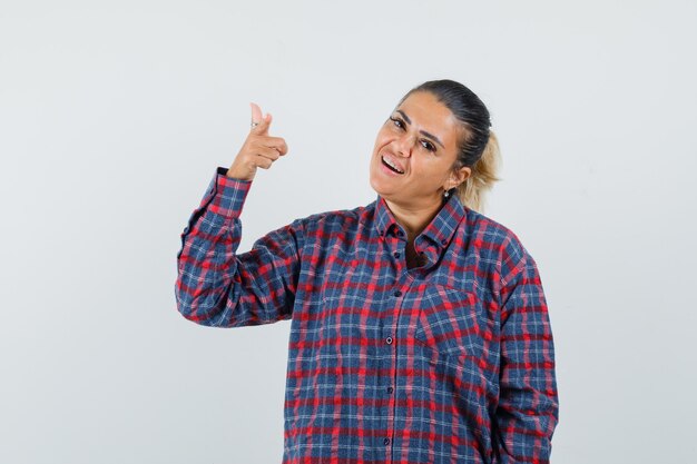 Young woman pointing at camera in checked shirt and looking happy , front view.