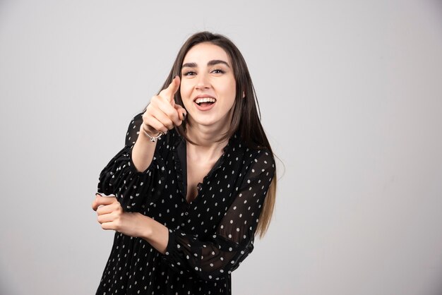 Una giovane donna che punta alla telecamera su un muro grigio.