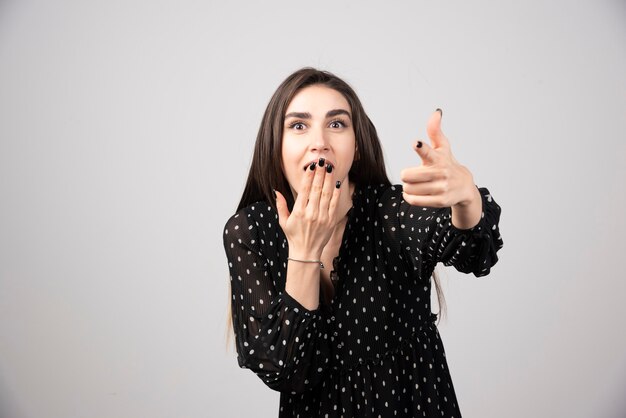 A young woman pointing to the camera on a gray wall.