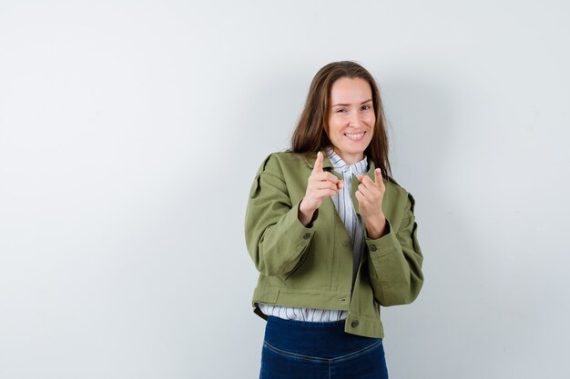 Young woman pointing at camera in shirt, jacket and looking glad, front view.