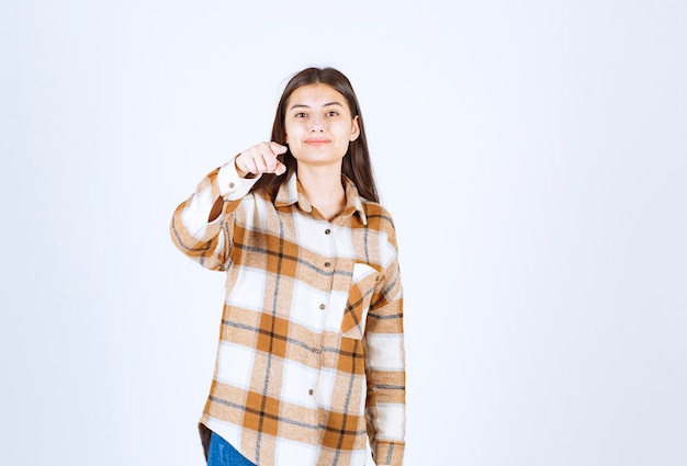 young woman pointing at camera on white wall. 