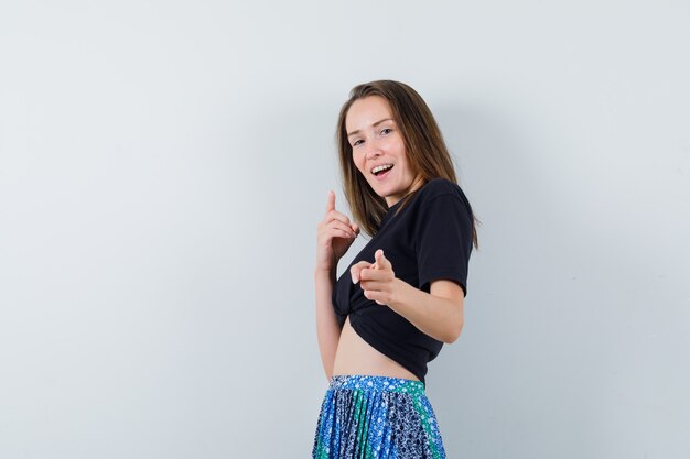 Young woman pointing at camera with index fingers in black t-shirt and blue skirt and looking happy