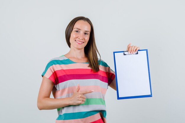 young woman pointing at clipboard in t-shirt and looking cheerful. front view.