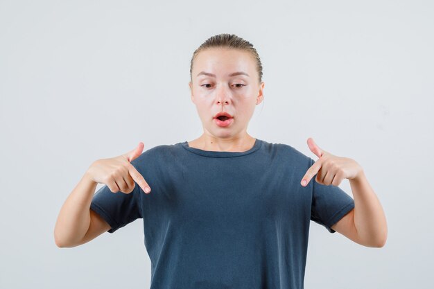 Young woman pointing down in grey t-shirt and looking surprised