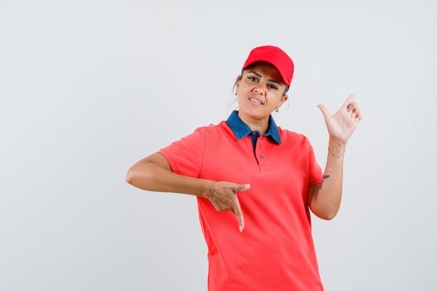 Young woman pointing down and right with fingers in red shirt and cap and looking pretty. front view.