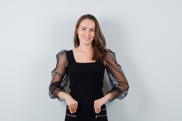 Young woman pointing down with both hands in black blouse and black pants and looking happy. front view.