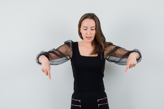 Young woman pointing down with both index fingers and looking down in black blouse and black pants and looking optimistic 