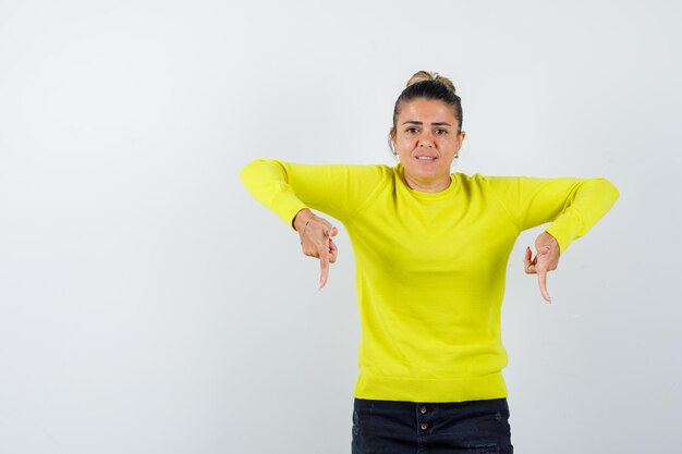 Young woman pointing down with index fingers in yellow sweater and black pants and looking harried 