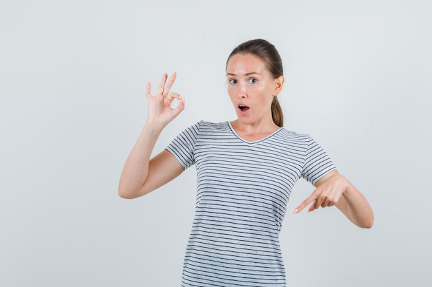 Young woman pointing down with ok sign in t-shirt and looking amazed , front view.