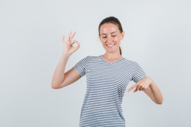 Young woman pointing down with ok sign in t-shirt and looking glad. front view.