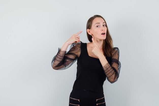 Young woman pointing to ear in black blouse and black pants and looking focused , front view.