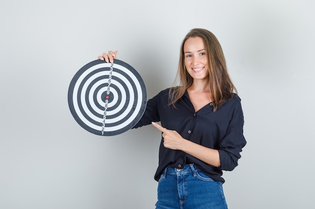 Foto gratuita giovane donna che punta il dito al bersaglio in camicia nera, pantaloncini di jeans e sembra allegro