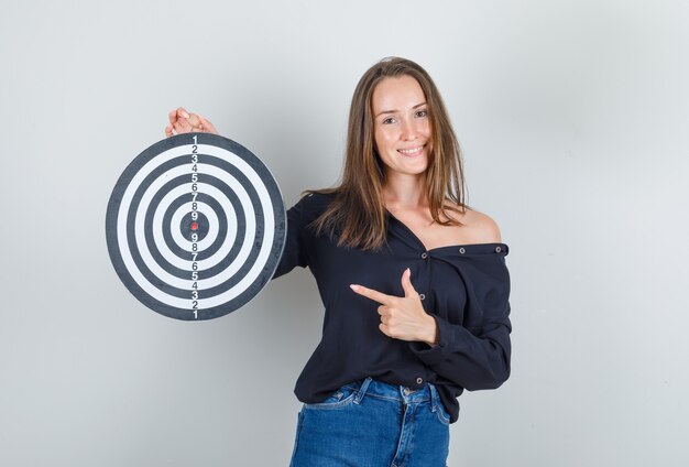 Young woman pointing finger at dartboard in black shirt, jeans shorts and looking merry