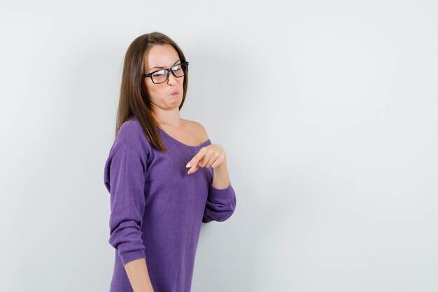 Young woman pointing finger down in violet shirt and looking disgusted. front view.