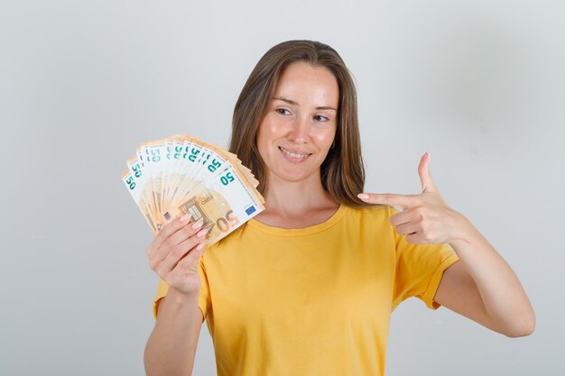 Young woman pointing finger at euro banknotes in yellow t-shirt and looking happy