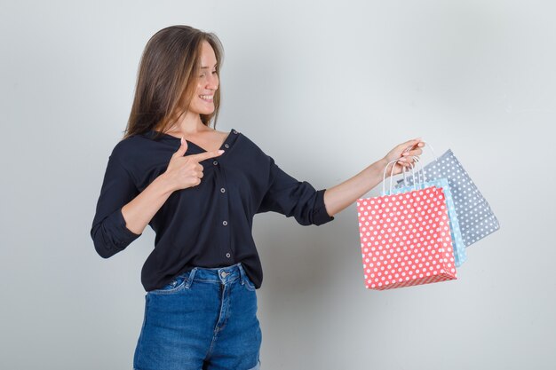 Young woman pointing finger at paper bags in black shirt, jeans shorts and looking joyful