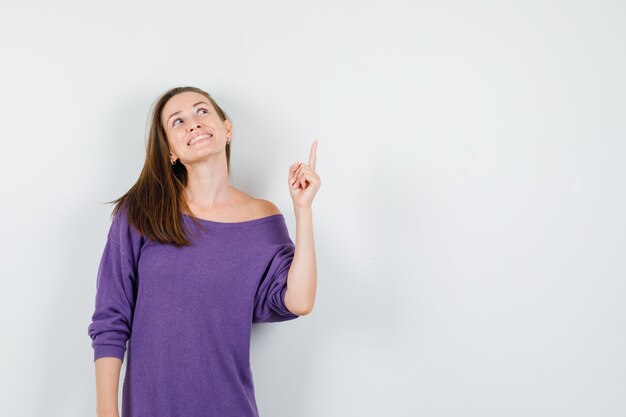 Молодая женщина указывая пальцем вверх в фиолетовой рубашке и глядя обнадеживающий, вид спереди.