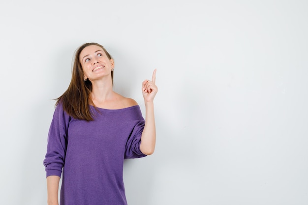 Young woman pointing finger up in violet shirt and looking hopeful , front view.