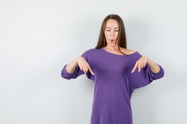 Young woman pointing fingers down in violet shirt and looking amazed , front view.