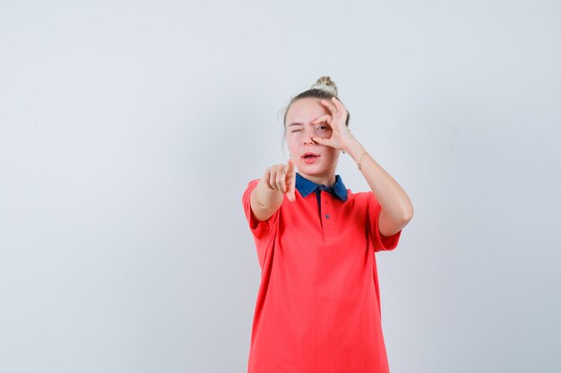 Young woman pointing at front, looking through fingers in t-shirt