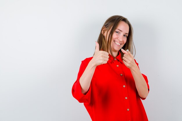 Young woman pointing at front, showing thumb up in red blouse and looking attractive , front view.