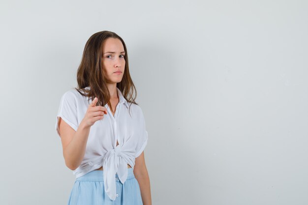 Young woman pointing at front with index finger in white blouse and light blue skirt and looking serious