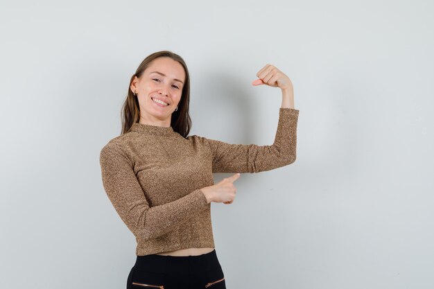 Young woman pointing to her arm muscle in golden blouse and looking confident , front view.