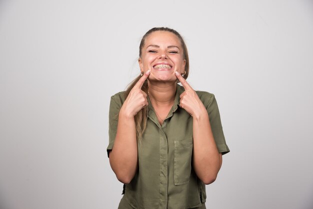 Young woman pointing at her cheek on gray wall.