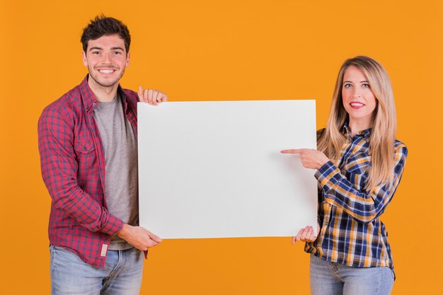 Young woman pointing her finger on placard hold by his boyfriend against orange background