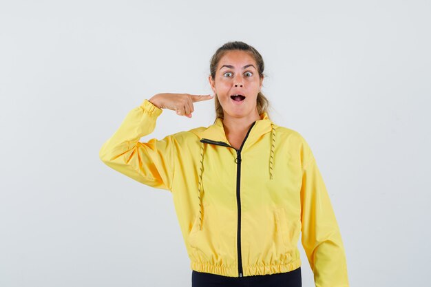 Young woman pointing at her opened mouth in yellow raincoat front view.