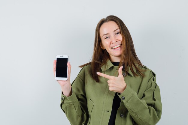 Young woman pointing at her phone in green jacket and looking satisfied , front view.