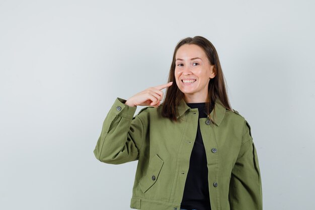 Young woman pointing at her teeth in green jacket front view.