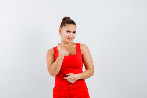 Young woman pointing at herself in red tank top, pants and looking curious , front view.