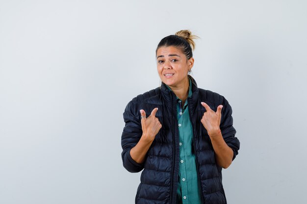 Young woman pointing at herself in shirt, puffer jacket and looking merry , front view.