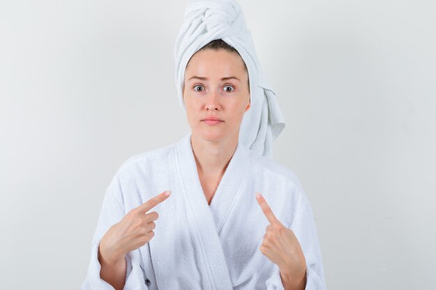 Молодая женщина, указывая на себя в белом халате, полотенце и озадаченно, вид спереди.