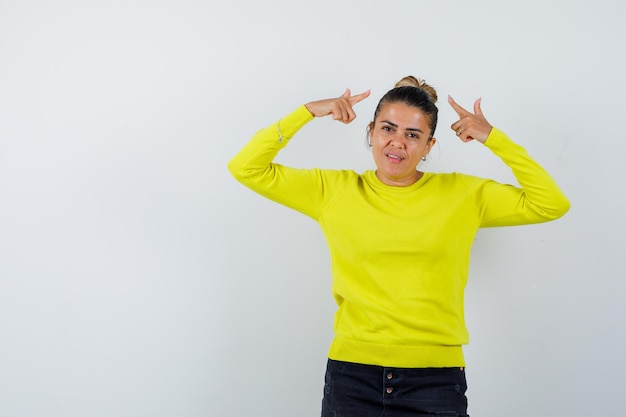 Young woman pointing at herself with index fingers in yellow sweater and black pants and looking serious