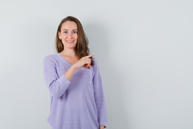 Young woman pointing at left in lilac blouse and looking glad 