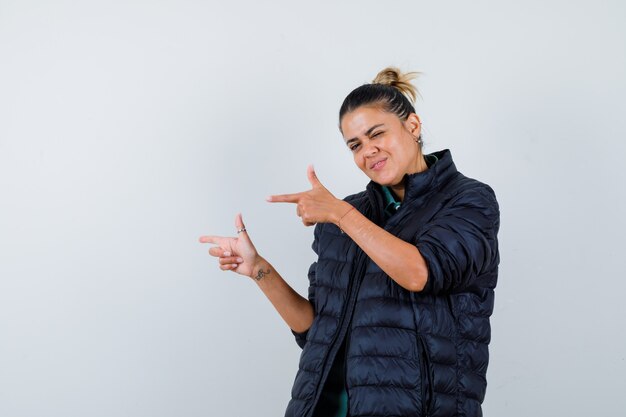 Young woman pointing left in puffer jacket and looking cheery. front view.