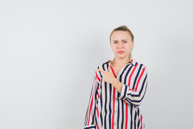 Young woman pointing left in striped blouse and looking confused