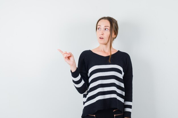 Young woman pointing left in striped knitwear and black pants and looking pensive