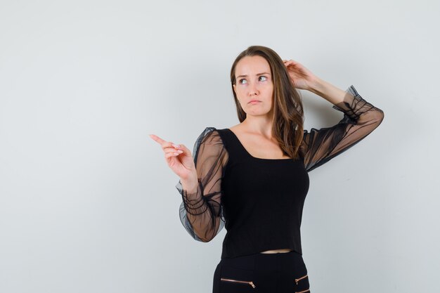 Young woman pointing left and thinking about something in black blouse and black pants and looking pensive. front view.