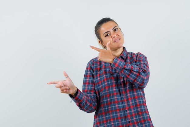 Young woman pointing left with both index fingers in checked shirt and looking happy. front view.