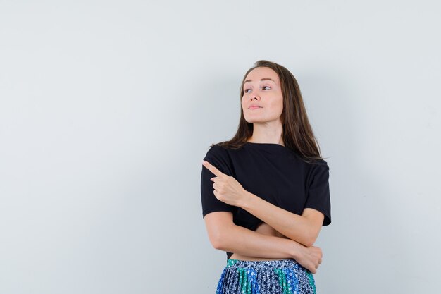 Young woman pointing left with index finger in black t-shirt and blue skirt and looking happy