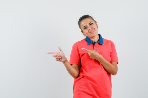 Молодая женщина, указывая влево указательными пальцами, улыбается в красной футболке и выглядит красивой, вид спереди.