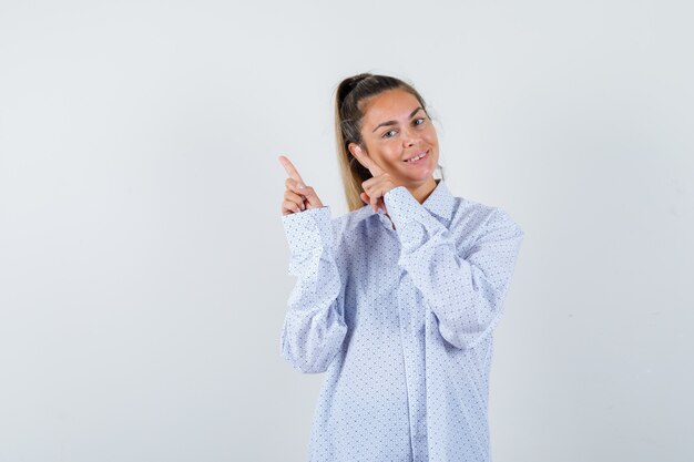 Молодая женщина указывает влево указательными пальцами в белой рубашке и выглядит счастливой