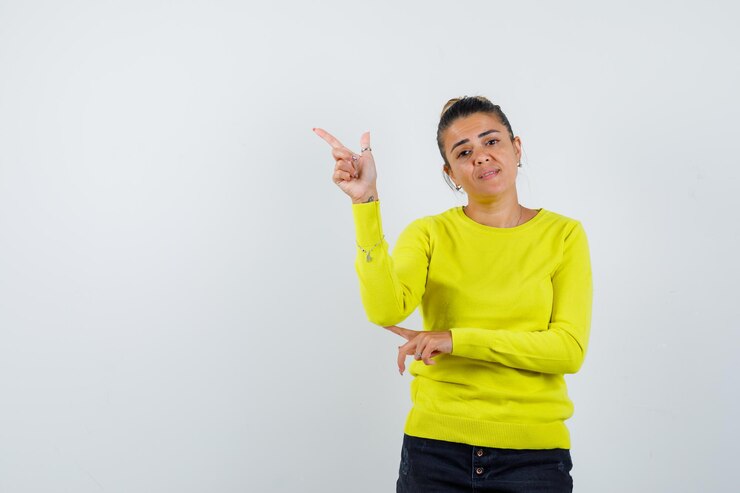 मुफ़्त फोटो young woman pointing left with index fingers in yellow sweater and black pants and looking happy 