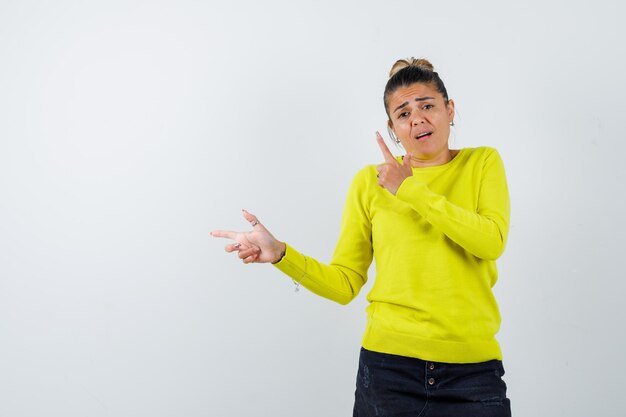 Young woman pointing left with index fingers in yellow sweater and black pants and looking happy