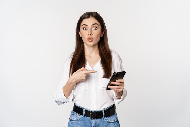 Young woman pointing at mobile phone while looking interested in app, showing smth on smartphone, standing over white background.