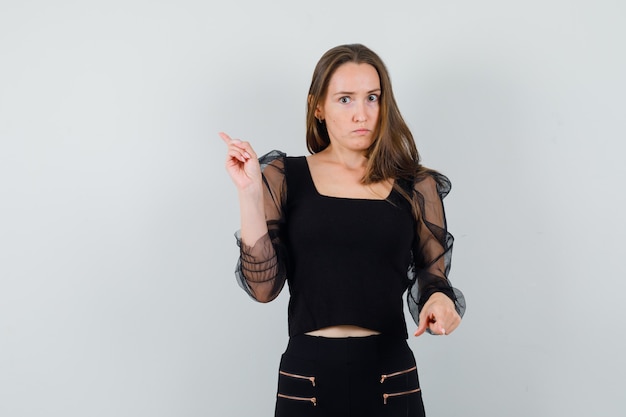 Young woman pointing opposite directions with index fingers in black blouse and black pants and looking serious. front view.