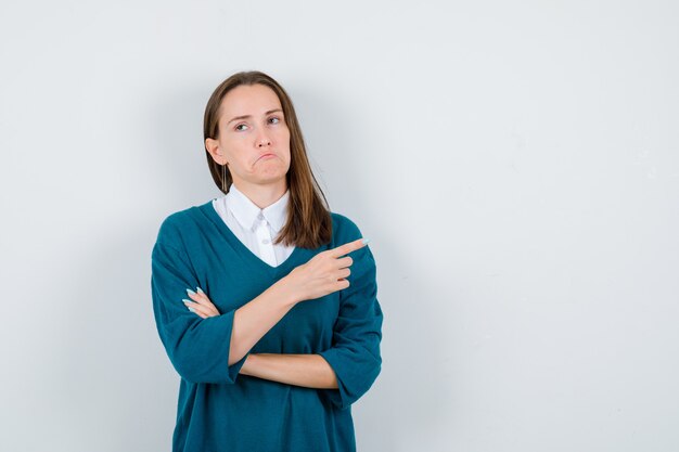 Молодая женщина указывает вправо, смотрит в сторону, изгибает нижнюю губу в свитере над белой рубашкой и выглядит невежественной, вид спереди.